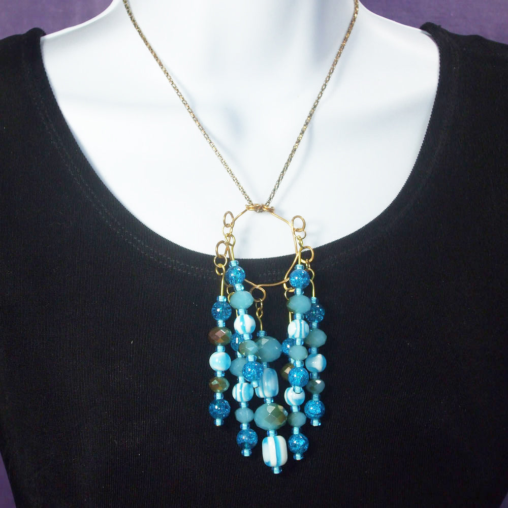 7 strands of various styles, cuts, shapes and textures of blue glass beads dangle on bronze wire from circular bronze wire design pendant.  Each strand giving a flash of color and blitz to this elegant necklace.